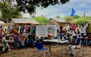 Man sits at a table in front of a sign with a crowd sitting around him in Mozambique