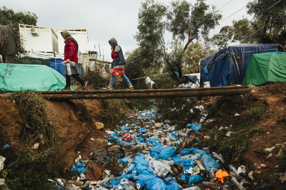 Two people cross a bridge over a ditch filled with litter on Aegean islands