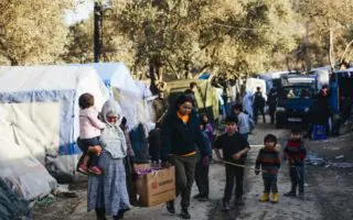 Family of refugees carry a box among tents and a dirt road on Greek islands