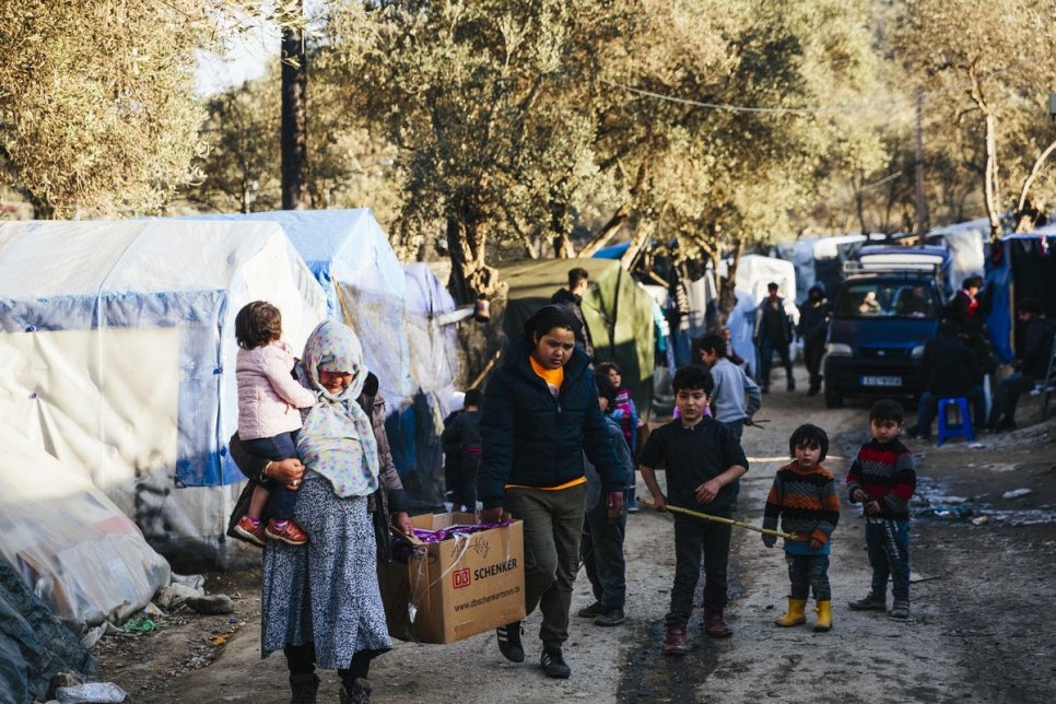 Family of refugees carry a box among tents and a dirt road on Greek islands