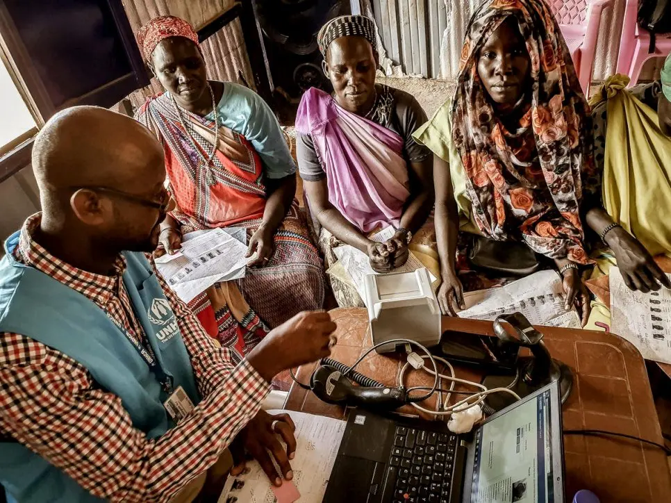 Man in a UNHCR blue vest sits at a computer and registers 4 women from South Sudan