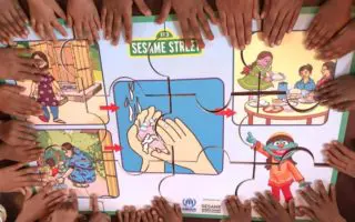 A circle of children's hands touching a puzzle advocating for hand washing with the Sesame Street logo on it