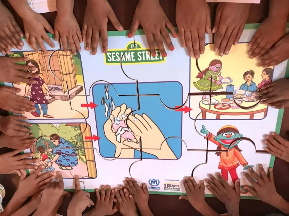 A circle of children's hands touching a puzzle advocating for hand washing with the Sesame Street logo on it