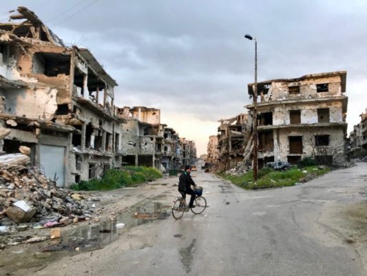 A man on a bicycle rides through a ruined street in Syria