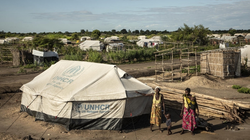 Two women and a child standing beside a tent that has the UNHCR logo on it
