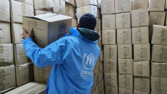 A man wearing a UNHCR jacket stacks boxes of medical supplies in the midst of