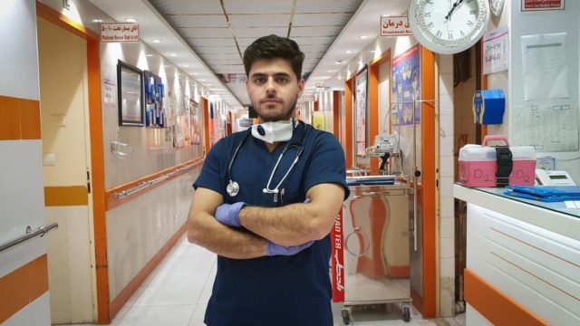 A male nurse stands in a hallway with his arms crossed