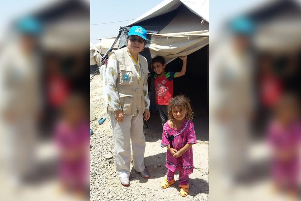 Woman wearing UNHCR branded clothing stands beside children at the entrance of a canvas tent