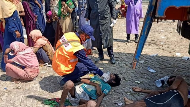 A man in a visibility vest gives water to a man who has collapsed on the ground