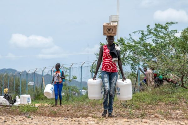 A woman balances a box on her head as she carries two jugs of water in both hands