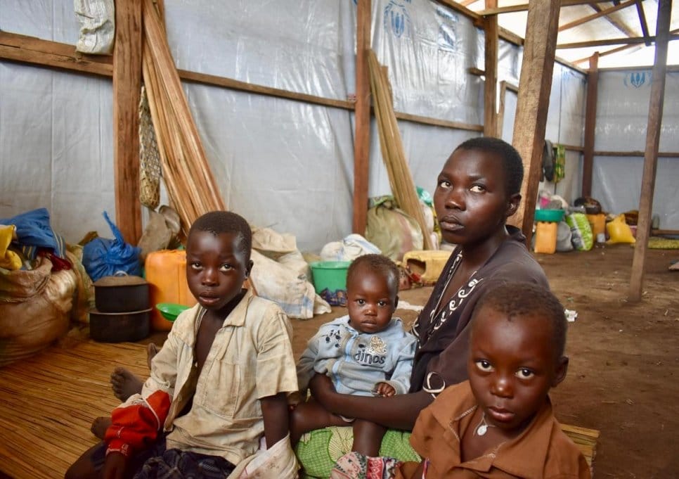 A woman sits on the floor in a makeshift house surrounded by her three children