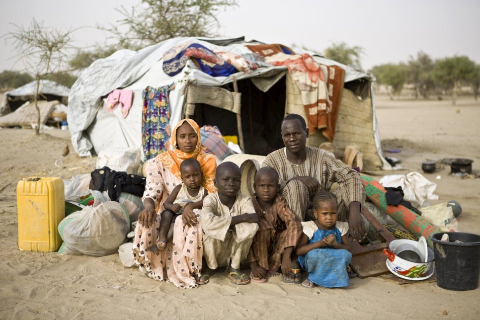 A family huddles together while sitting on the ground in front of a makeshift tent