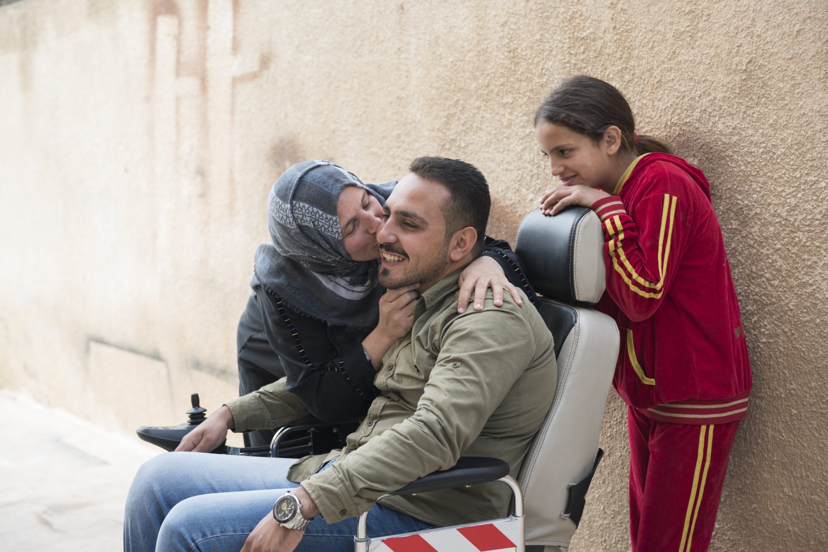 A woman gives a man in a wheelchair a kiss on the cheek while a young girl hangs off the back of the wheelchair