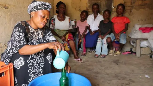 Justine Mbilizi pours palm wine into recycled glass bottles next to family.