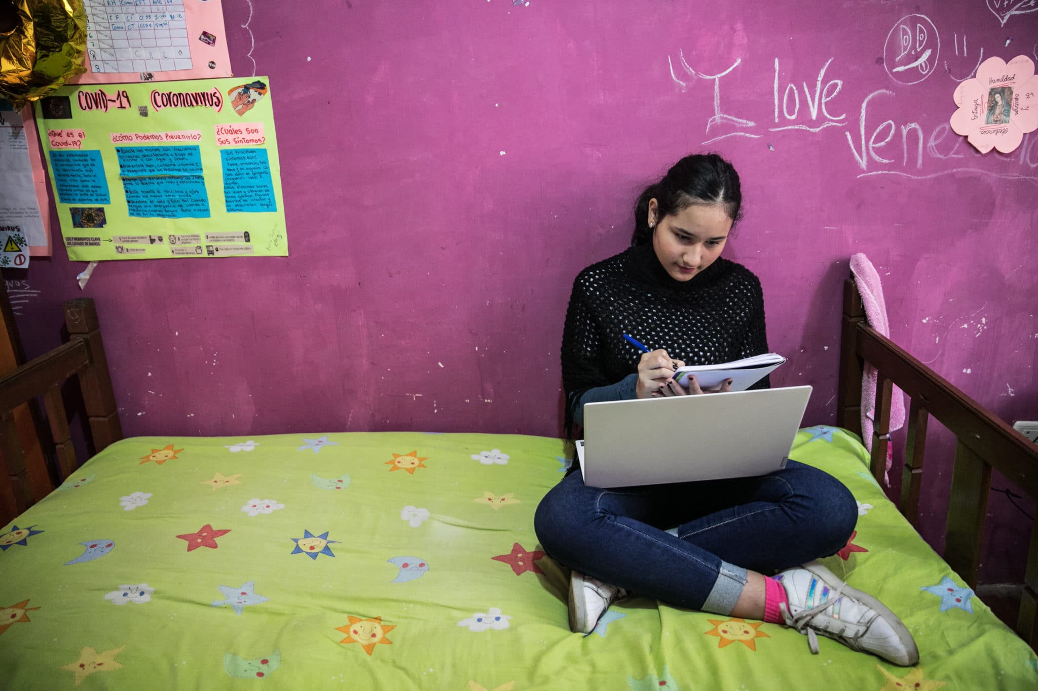 Teenage girl with dark hair pulled back into a ponytail sits crosslegged on her bed. She is wearing a black turtleneck, jeans and running shoes. She is holding a pen in her right hand writing in a notebook with a laptop computer in her lap.