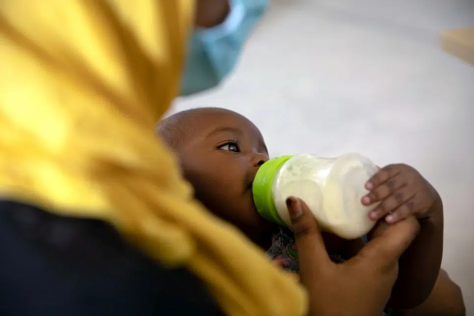 Baby holding bottle being fed by refugee mother wearing yellow headscarf