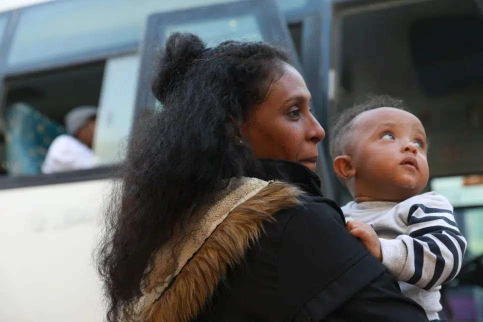 Woman in black fur-trimmed hooded jacket holds baby in front of bus