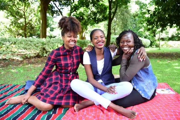 Three women smiling and sitting on a red patterned picnic blanket in front of a green foliage backdrop. Their arms are draped across each other's shoulders.