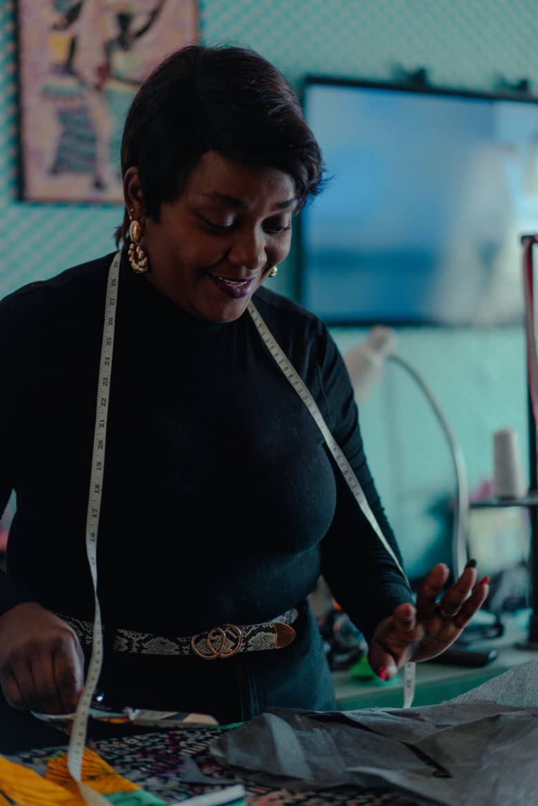 Dark skinned woman with a short pixie cut wearing black long sleeved turtle neck shirt and bold gold earrings, has measuring tape around her neck is looking down at her fabric.