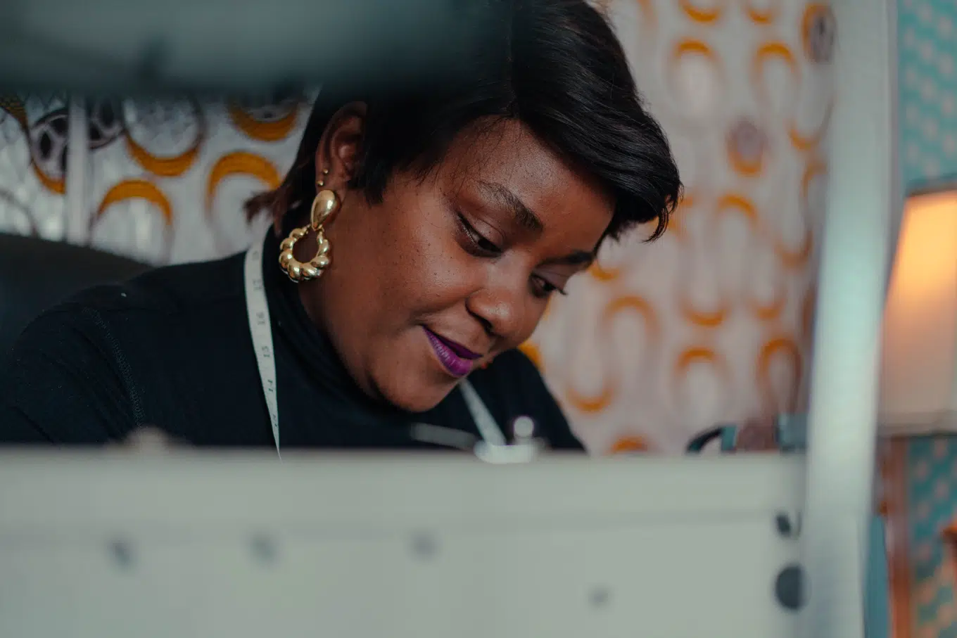 Dark skinned woman with a short pixie cut wearing black long sleeved turtle neck shirt and bold gold earrings, sits at her work station with a measuring tape around her neck. Her face in the image is framed by a sewing machine in the front.