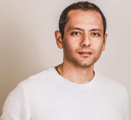 Portrait photo of man in white long-sleeved shirt standing in front of beige wall.