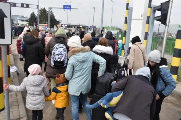 Group of people of varying ages and gender walking towards border crossing with their suitcases and bags.