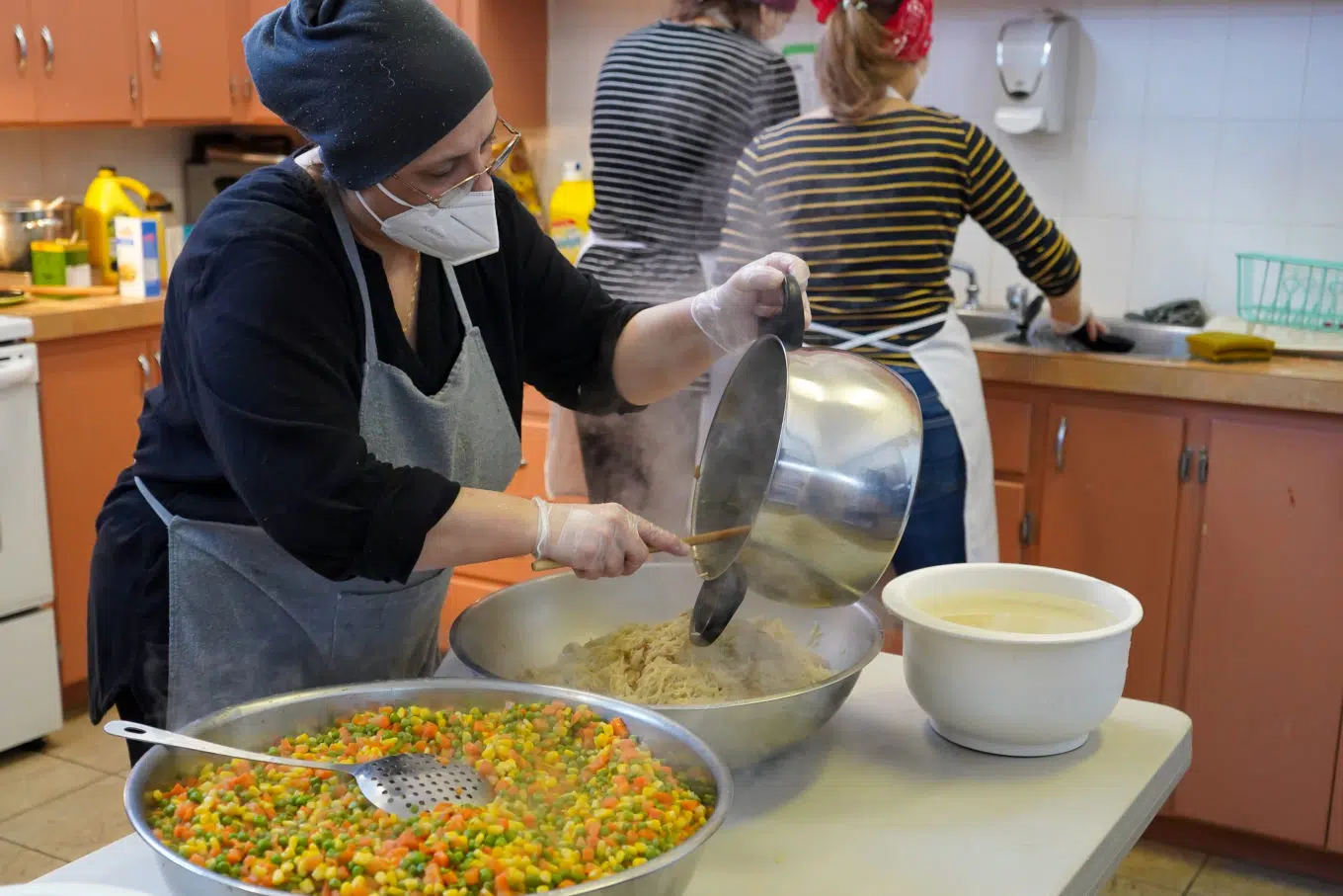 Woman in black shirt and grey apron holds a pot with rice. She is scooping the rice into a larger bowl. There is also a bowl of carrots and peas next to her.