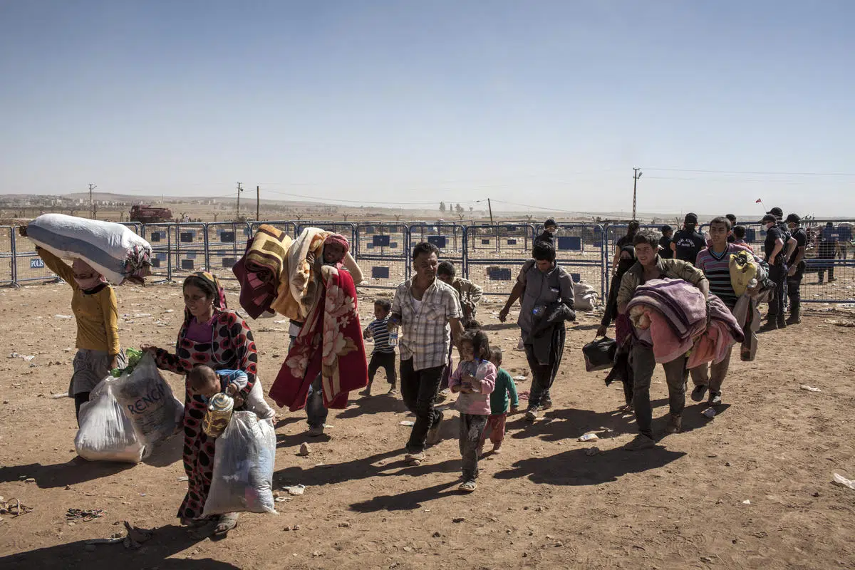 Refugees walking on dirt plane in front of metal fencecarrying belongings