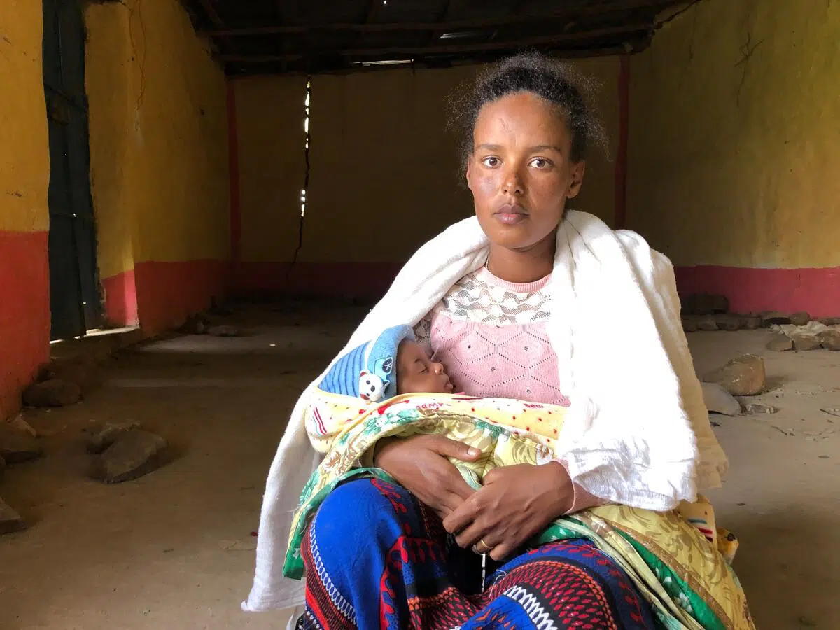 Ethiopian woman sitting holding her sleeping baby in an empty concrete room with yellow and red walls and pieces of rubble behind her