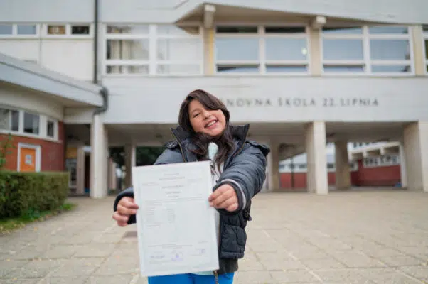 Young girl standing at school entrance excitedly holding her report card out in front of her