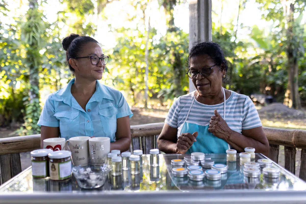 two women standing behind a glass table with various products on display, behind them is an opening looking out onto a