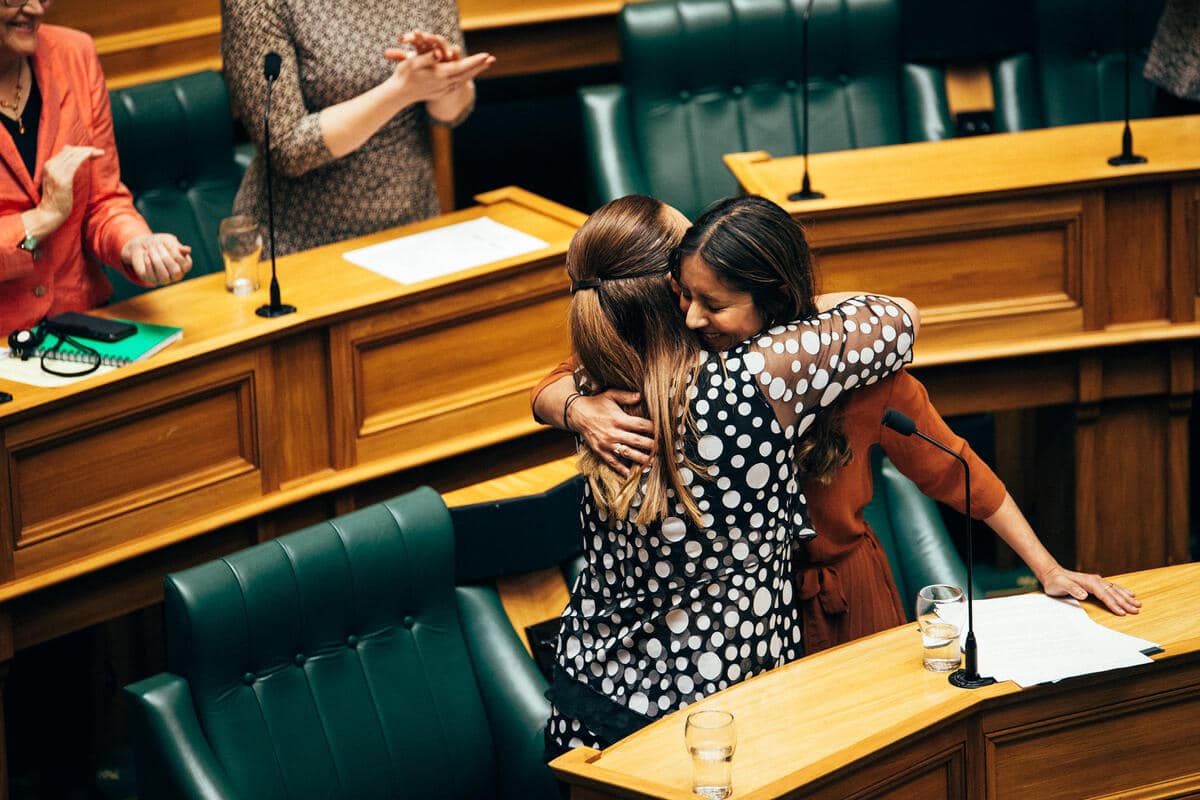 Two women hugging behind desks with green leather chairs in parliamentary style room