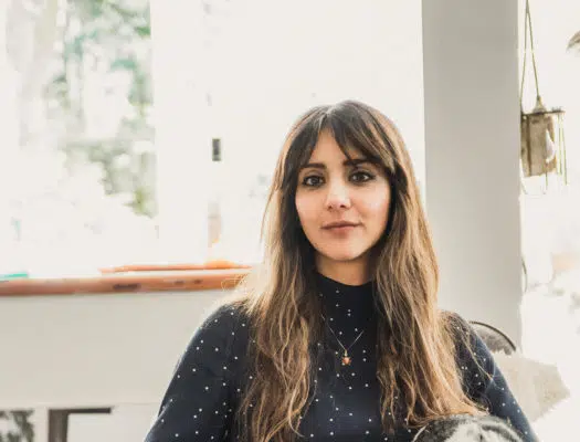 Iranian woman with long brown hair and bangs sitting in front of a window in a white room with bright backlighting