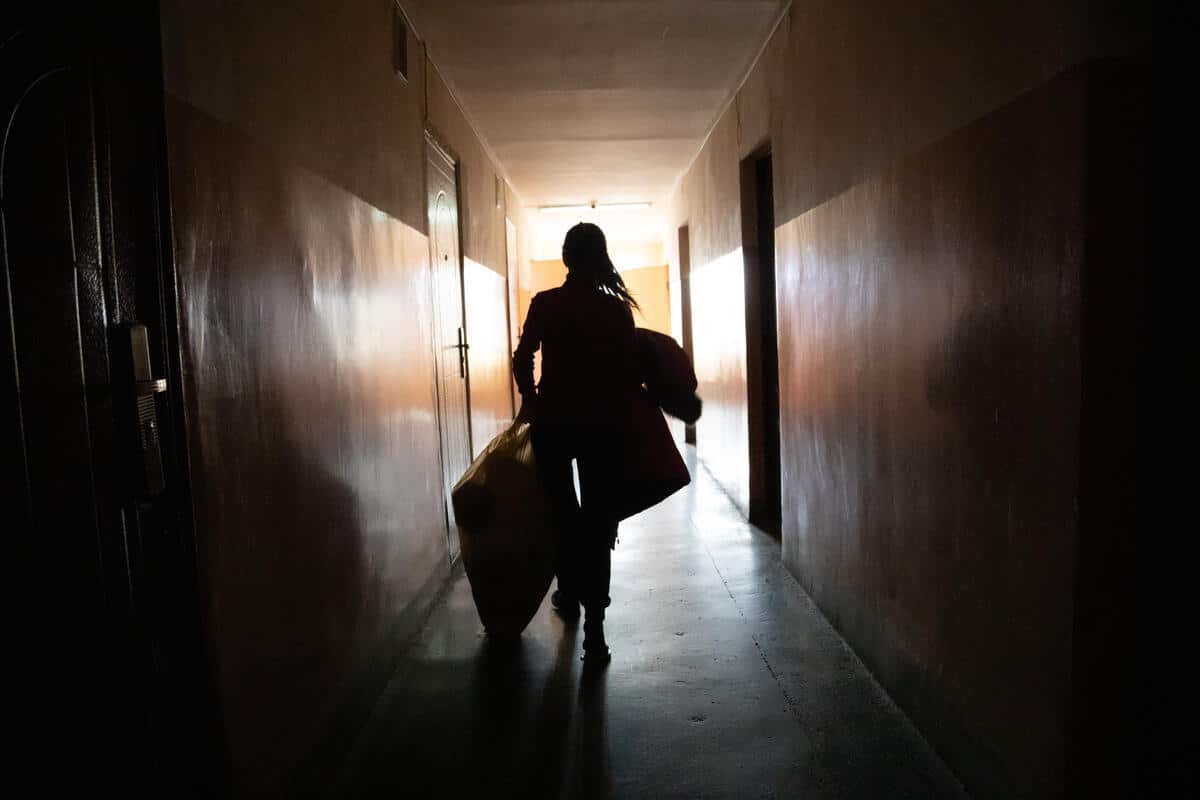 Young woman walking down a dark hallway carrying bags of clothes and blankets