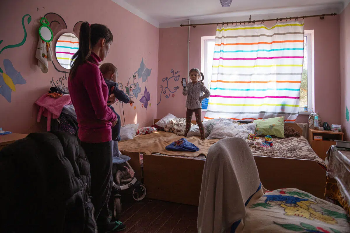 Woman holding a baby is standing in a pink room looking at a young female child, with her hair in pigtails, who is standing on a bed