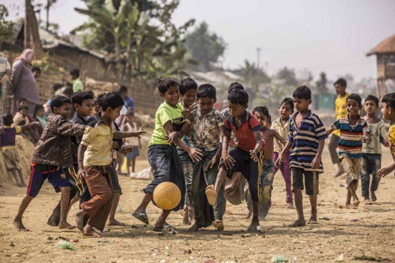 Many children play soccer barefoot on a dirt pitch in a refugee camp.
