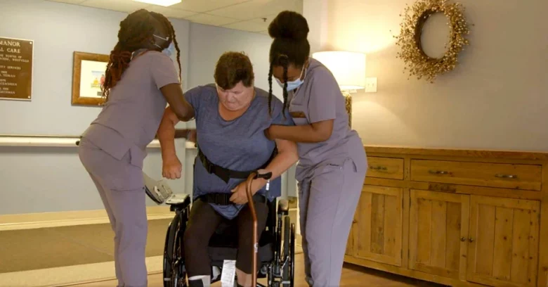 Two women in purple scrubs on both sides of a woman in a wheelchair helping her to stand up.