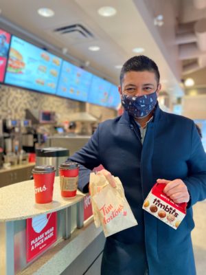 Man in blue peacoat hold donut box in his right hand that says timbits and a brown parchment bag that says Tim Hortons. He is leaning against a counter top with two red coffee cups. There are three screens in the back displaying Tim Horton's menu.