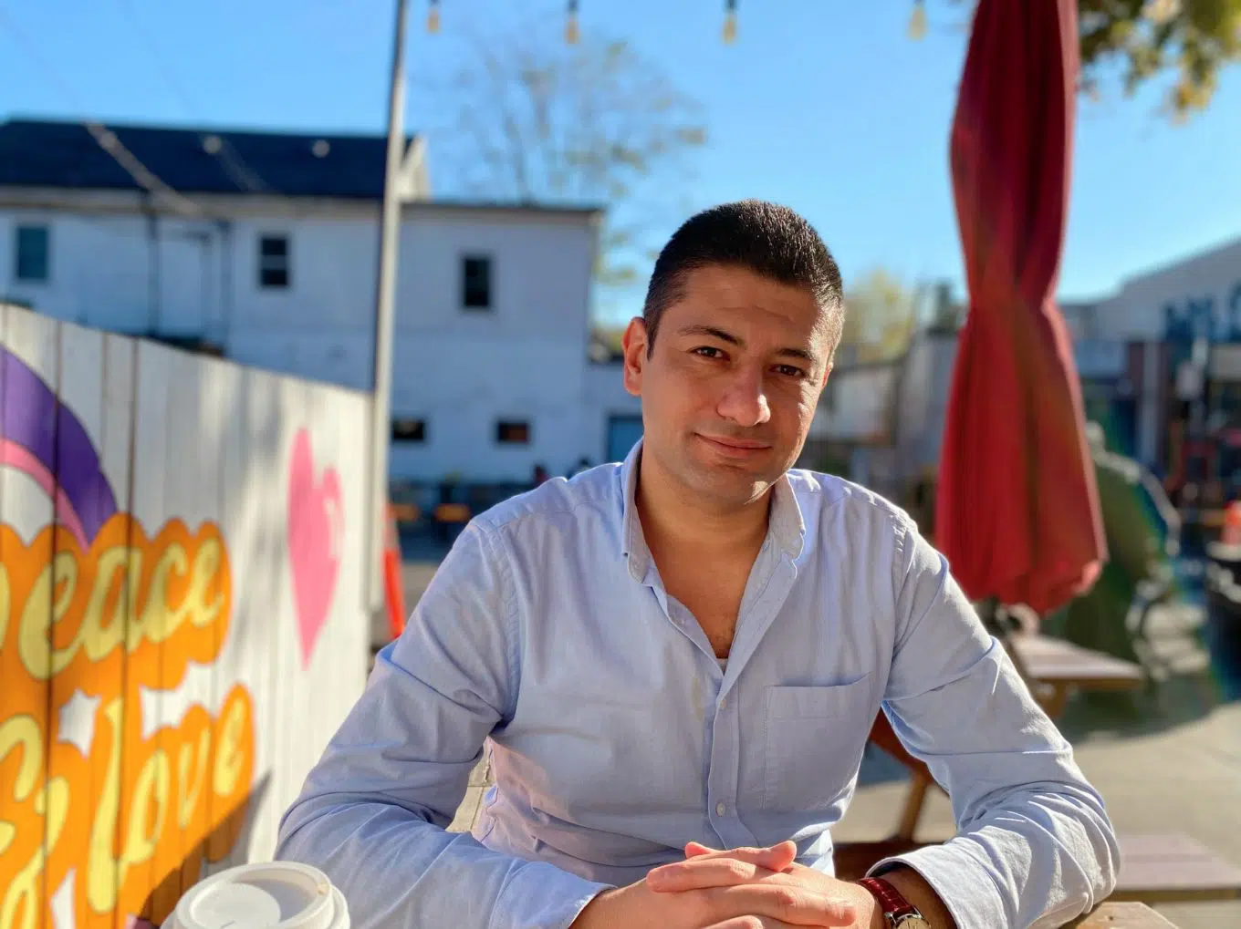 Man in light blue button up sits with hands clasped together. There is a closed red patio umbrella behind him to his right and a painted fence to his left.