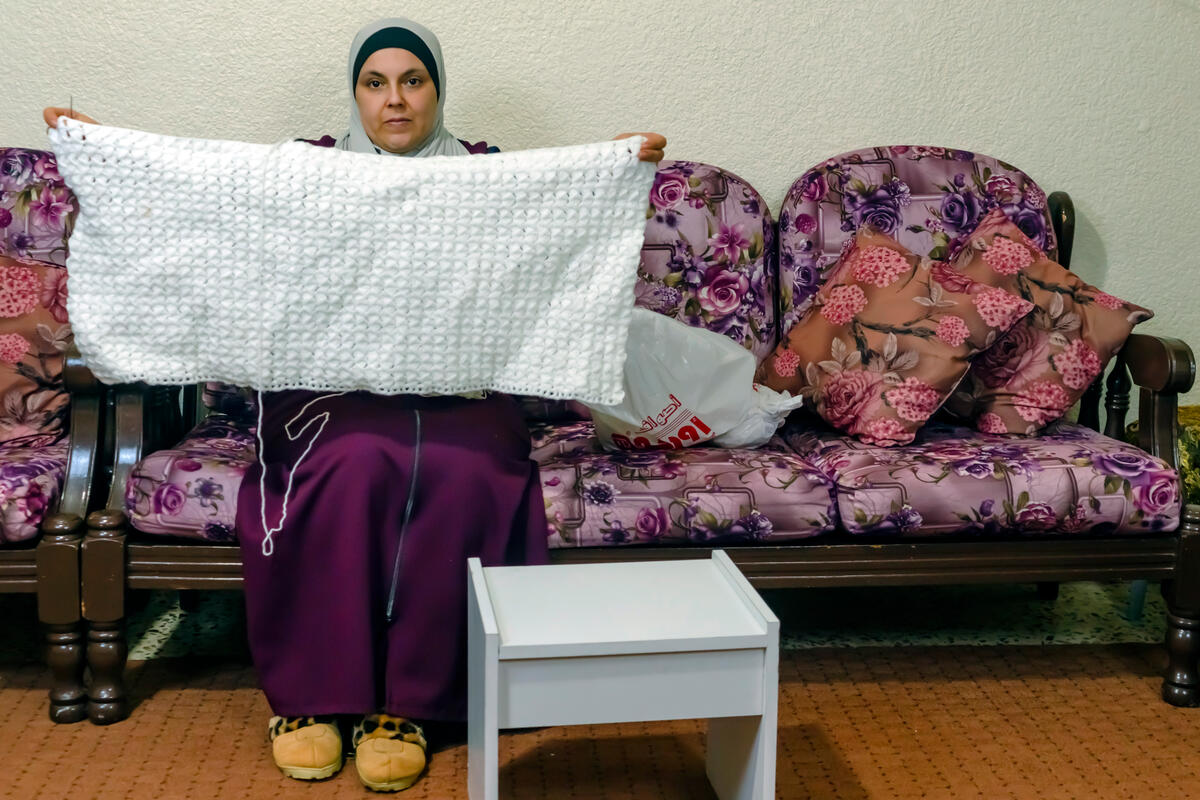 Woman wearing a hijab holding up a white blanket and sitting on a purple floral couch 