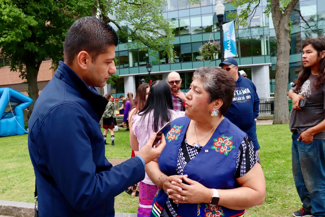 Man in blue peacoat stands to the left holding his phone in his hand next to a woman in a blue vest with floral beaded patterns on it.