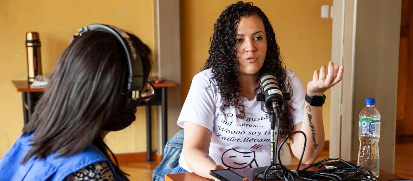UNHCR Worker wearing a blue vest with black hair and wearing headphones sitting with her back to the camera in front of a woman with curly black hair wearing a white t-shirt with writing on it, she is speaking into the microphone