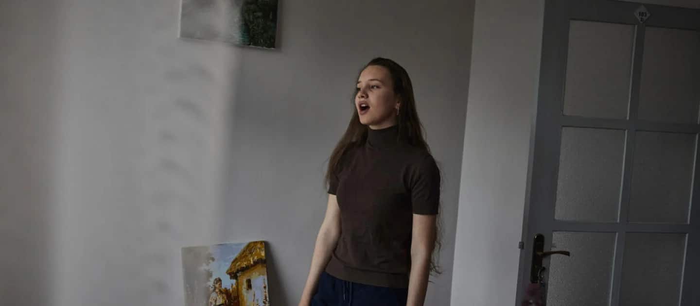 young woman with long straight brown hair wearing a brown short sleeve mock-neck shirt and black pants is singing with her arms at her side standing at a slight angle facing away from the camera