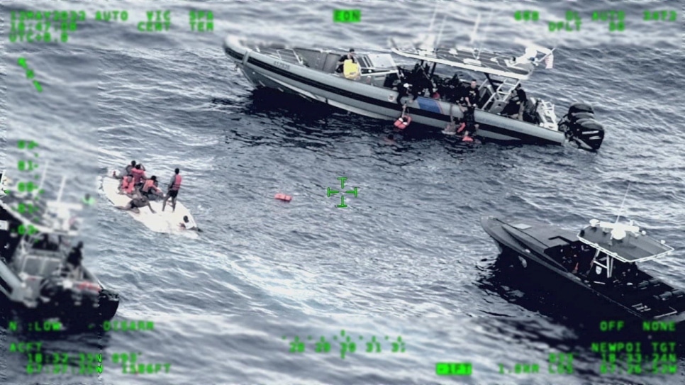 three rescue boats circles around a white smaller, capsized, boat with a group of people wearing life jackets sitting on top 