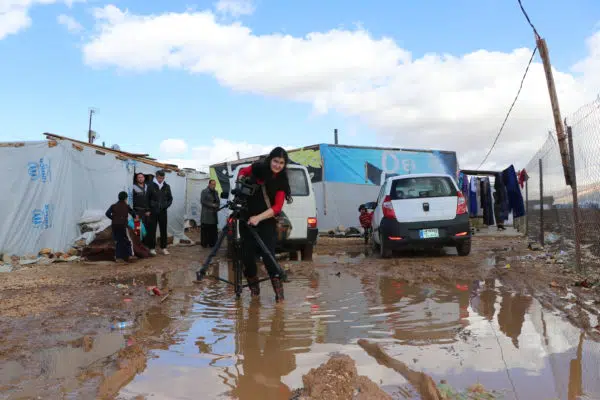 Woman wades through flooded ground with film camera on a tripod. In the background is a UNHCR shelter with people in the background.