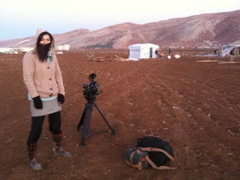 Woman in the middle of dark brown field with UNHCR shelter in the background. She has a film camera and a tripod next to her.