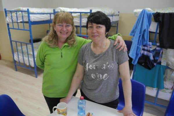 Two women, one in a green t-shirt with blonde shoulder length hair and the other with short black hair in a grey t-shirt, standing in front of a couple bunk beds