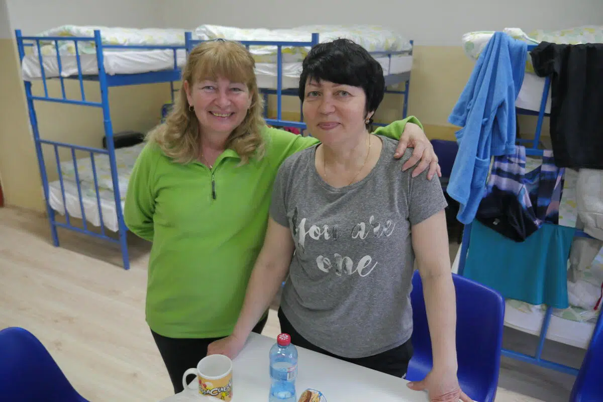 Two women, one in a green t-shirt with blonde shoulder length hair and the other with short black hair in a grey t-shirt, standing in front of a couple bunk beds