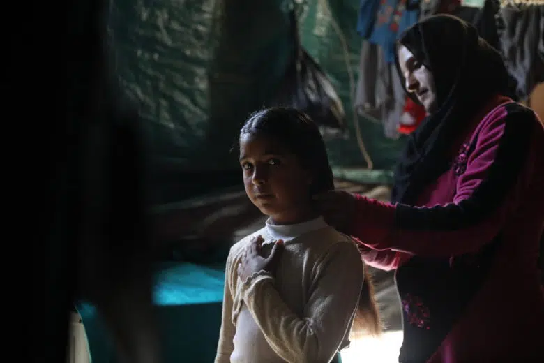 Young girl wearing white long-sleeve mock-neck shirt holds her hand across her chest and looks into the camera while a woman wearing a burgundy hijab and long-sleeve shirt brushes her hair