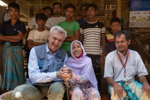 Filippo Grandi sitting next to and holding the hand of a smaller woman who is wearing a purple head scarf, there is a room of people standing behind them